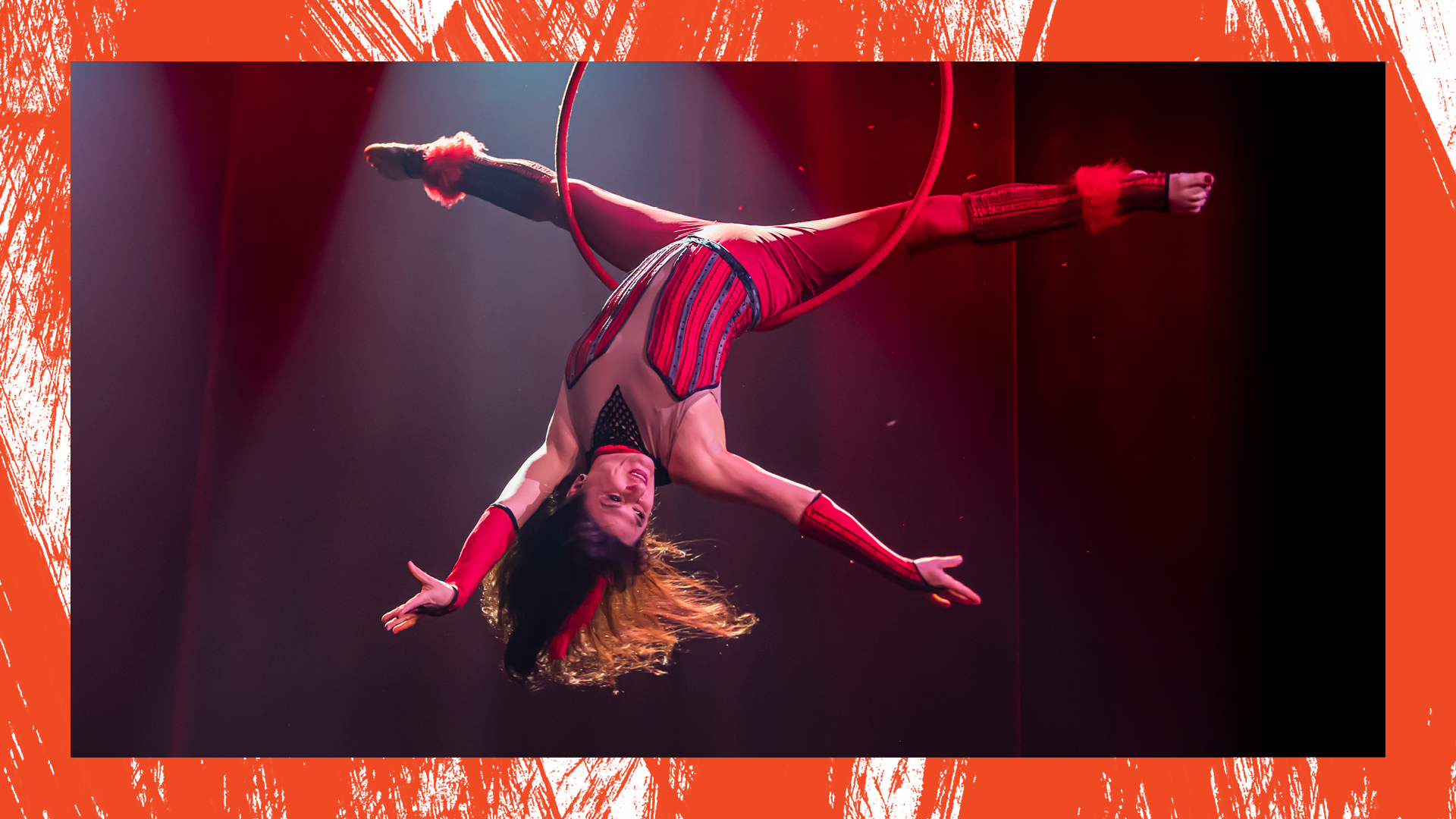 A woman wearing a red and black fitted costume is upside down inside of a hoop called a Lyra. She is balanced on the base of her spine, her legs in a full split as her head head hangs down and her long hair flows with movement.