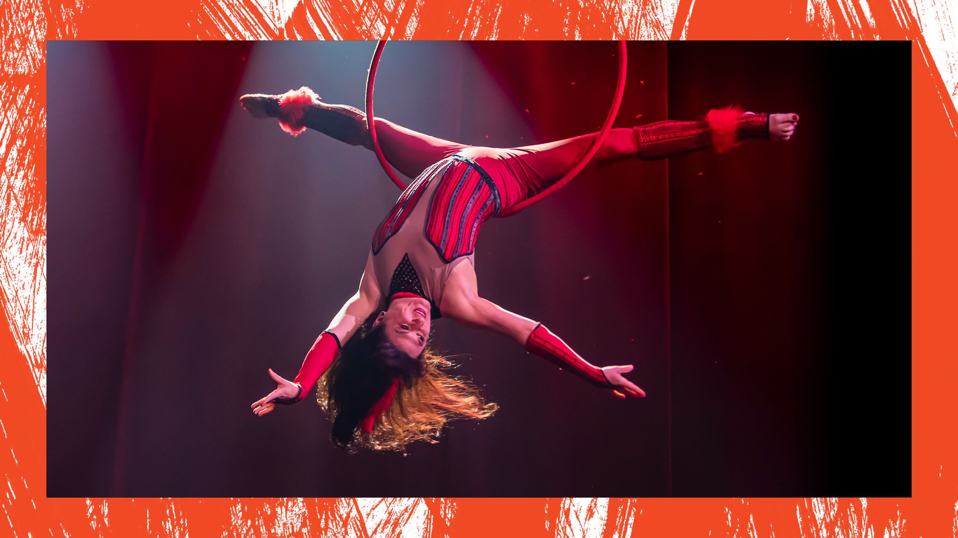 A woman wearing a red and black fitted costume is upside down inside of a hoop called a Lyra. She is balanced on the base of her spine, her legs in a full split as her head head hangs down and her long hair flows with movement.